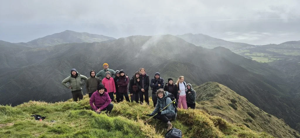 zdjęcie SKN Botaników Gatun na tle pięknej górskiej panoramy. Studenci są ubrani w górską odzież przeciwdeszczową. W tle majaczą pobliskie zalesione szczyty, jednak wszędzie panuje mgła. Na zdjęciu jest ok. 15 osób.